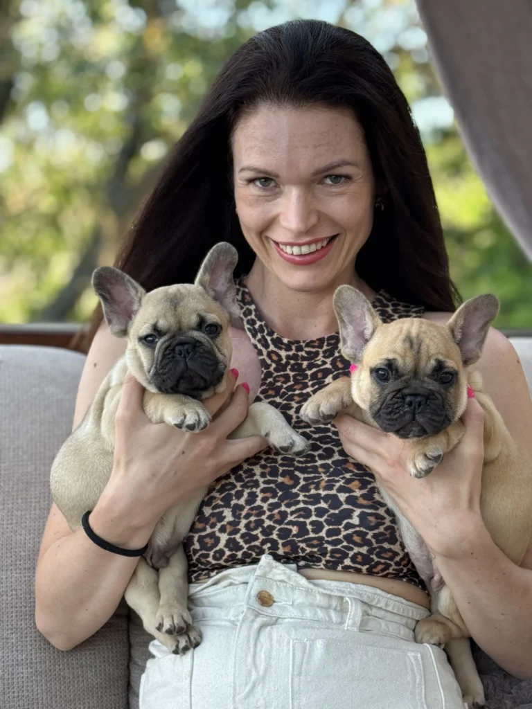 Woman smiling while holding two beige French Bulldog puppies outdoors.