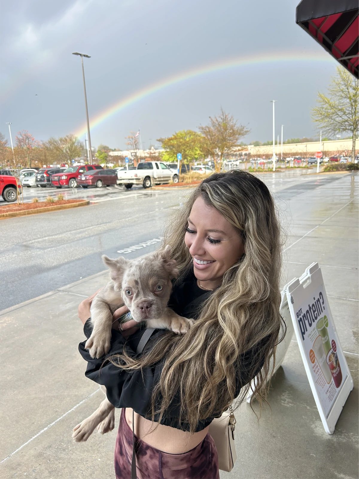 Woman holding a French Bulldog puppy with a rainbow in the sky behind her